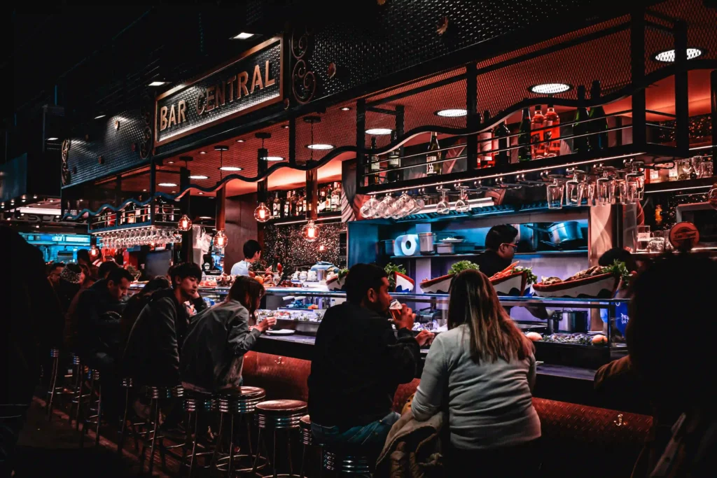 Busy bar counter with guests seated on stools at Bar Central, seafood display, hanging glasses and warm mood lighting