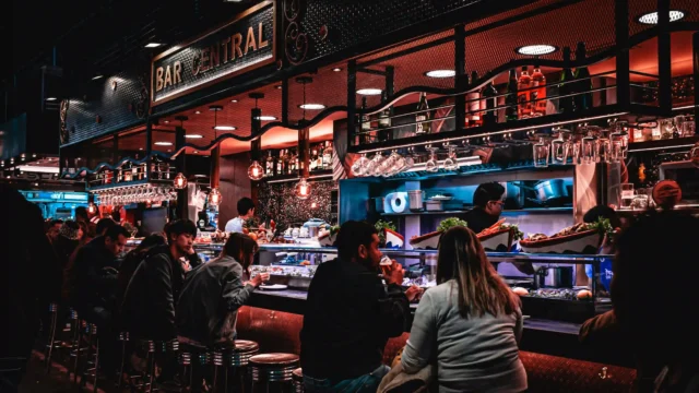 Busy bar counter with guests seated on stools at Bar Central, seafood display, hanging glasses and warm mood lighting