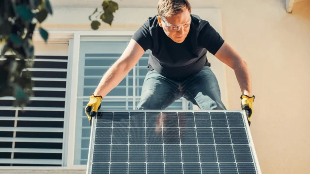 Technician wearing safety glasses and yellow gloves installing a solar panel on a residential rooftop