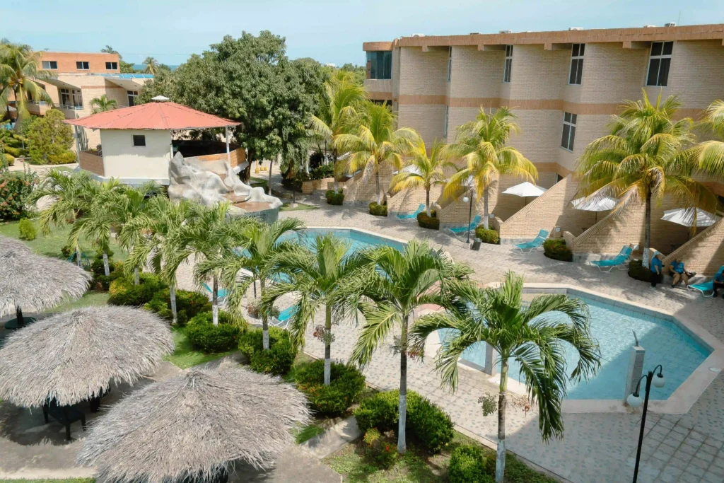 Aerial view of a tropical hotel resort with an outdoor pool, palm trees, sun loungers and thatched palapas