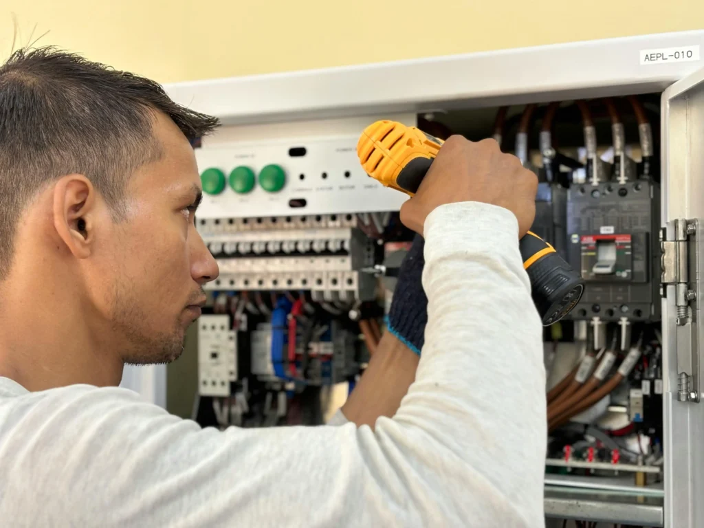 Electrician using a cordless drill to work on an electrical control panel with circuit breakers and wiring