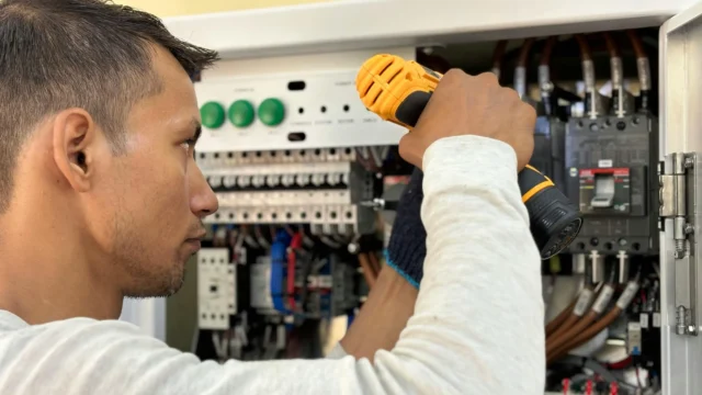 Electrician using a cordless drill to work on an electrical control panel with circuit breakers and wiring