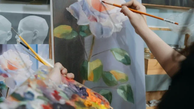 Artist painting a white rose in oil on canvas at an easel, holding a colorful palette in an art studio