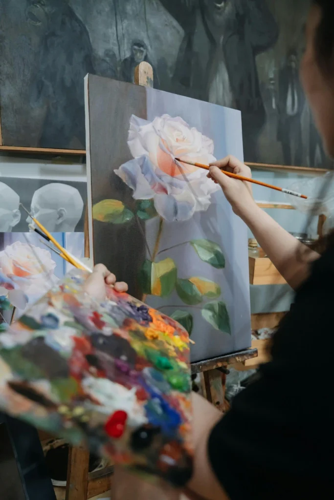 Artist painting a white rose in oil on canvas at an easel, holding a colorful palette in an art studio