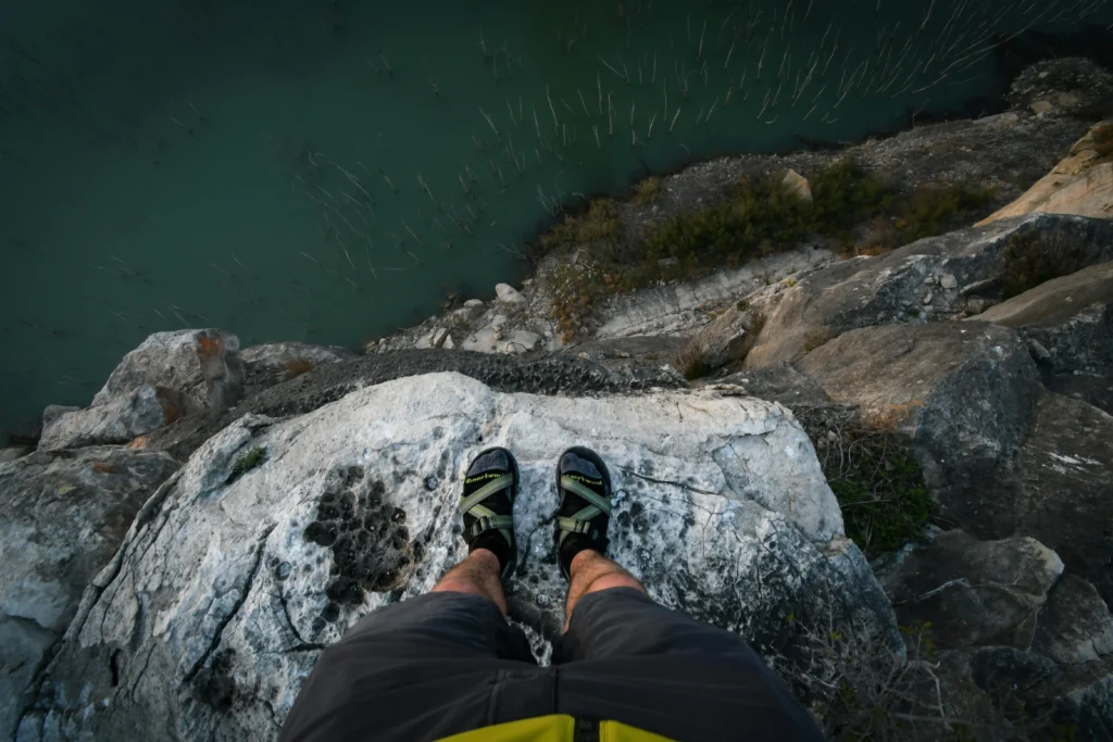 First-person view looking down from a rocky cliff over turquoise water, feet in sport sandals on the edge