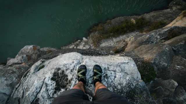 First-person view looking down from a rocky cliff over turquoise water, feet in sport sandals on the edge