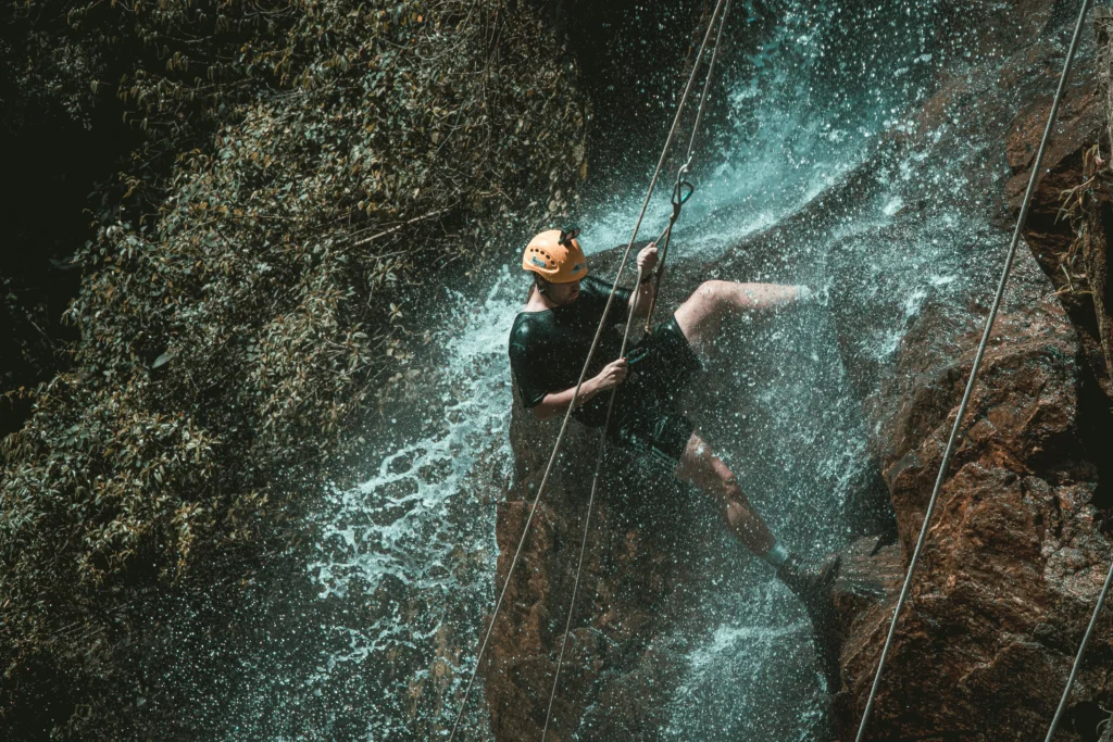 Person abseiling down a waterfall in a tropical canyon, wearing an orange safety helmet and harness