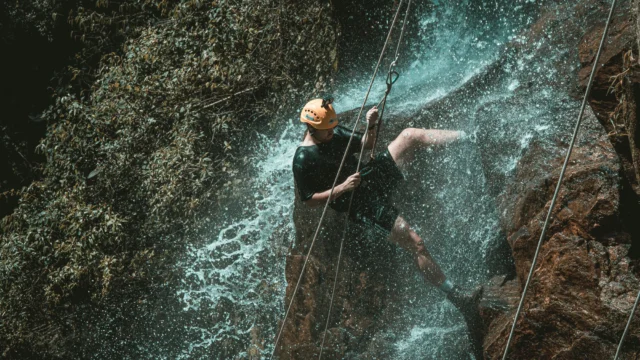 Person abseiling down a waterfall in a tropical canyon, wearing an orange safety helmet and harness