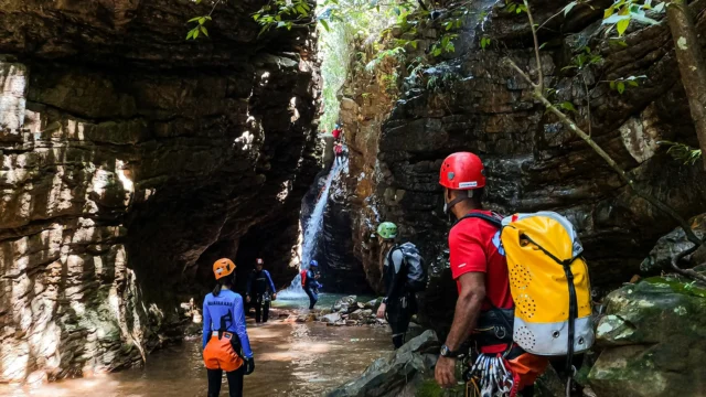 Group of canyoners with helmets and dry bags walking through a narrow rocky gorge toward a waterfall