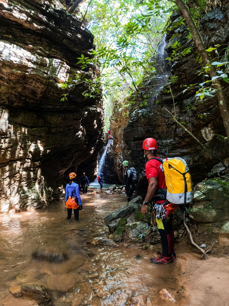 Group of canyoners with helmets and dry bags walking through a narrow rocky gorge toward a waterfall