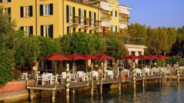 Elegant Italian lakeside hotel with yellow facade and waterfront terrace restaurant with red umbrellas and white chairs on a wooden dock