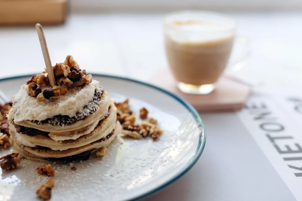 Stack of fluffy pancakes topped with walnuts, coconut flakes and chocolate on a plate next to a latte in a bright café setting