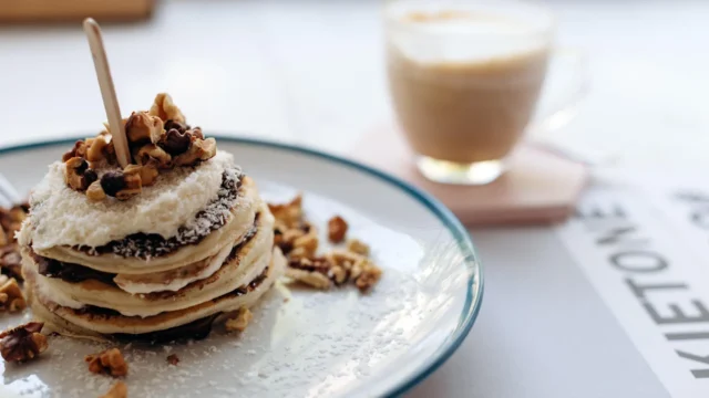 Stack of fluffy pancakes topped with walnuts, coconut flakes and chocolate on a plate next to a latte in a bright café setting