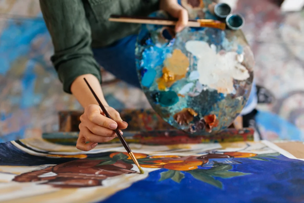 Close-up of an artist's hand painting a colorful floral canvas in blue and orange tones, holding a heavily used paint palette