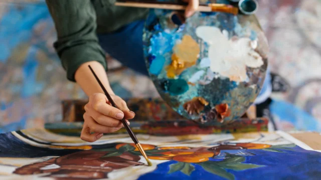 Close-up of an artist's hand painting a colorful floral canvas in blue and orange tones, holding a heavily used paint palette