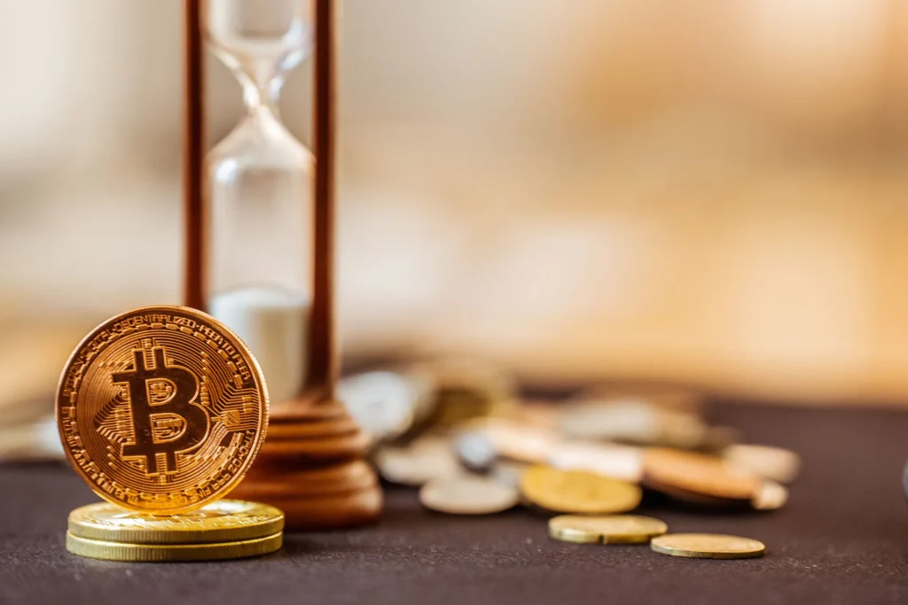 Golden Bitcoin coin leaning against a wooden hourglass with scattered coins on a dark surface, symbolizing time and value