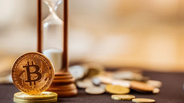 Golden Bitcoin coin leaning against a wooden hourglass with scattered coins on a dark surface, symbolizing time and value