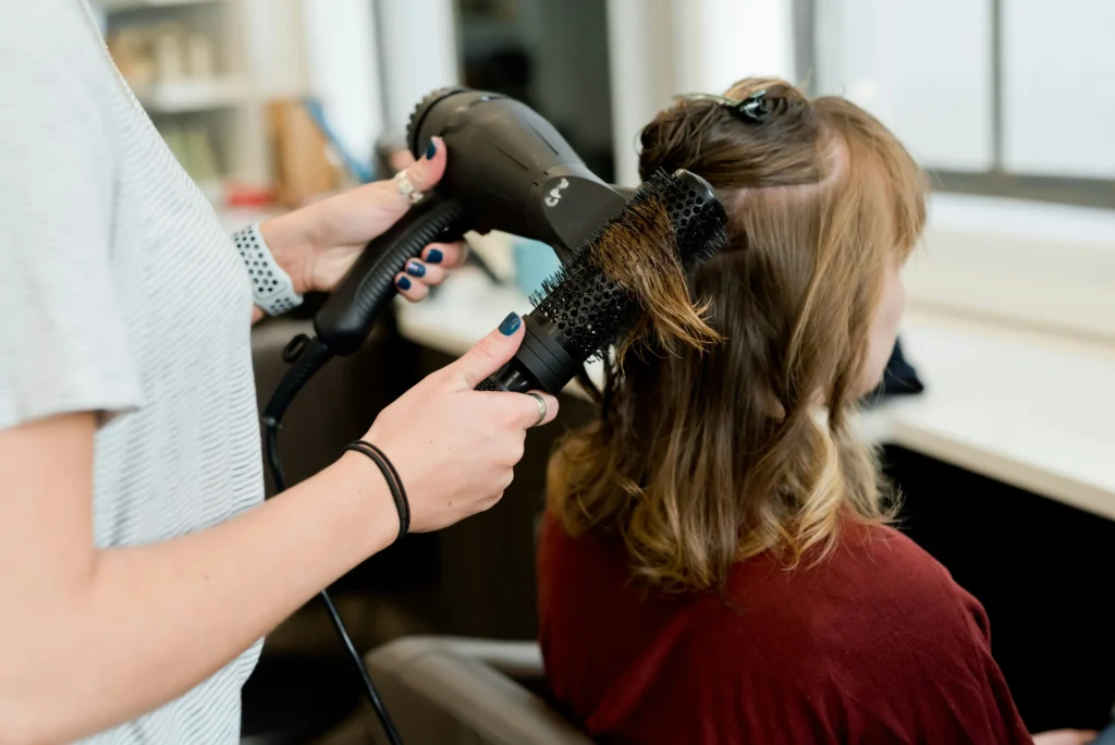 Hairdresser blow-drying and styling a client's hair with a round brush and hair dryer in a bright salon