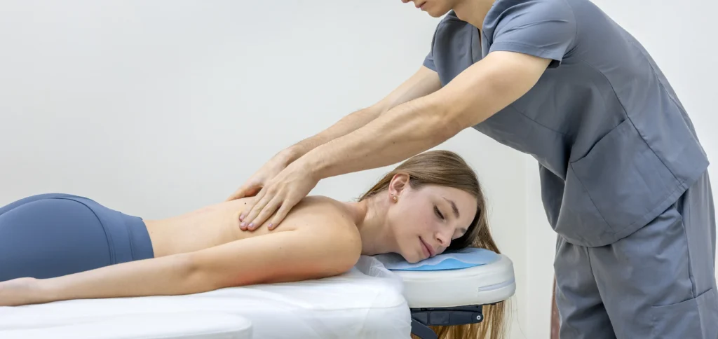 Therapist in grey scrubs performing a back massage on a relaxed female patient lying face down on a treatment table in a bright white room