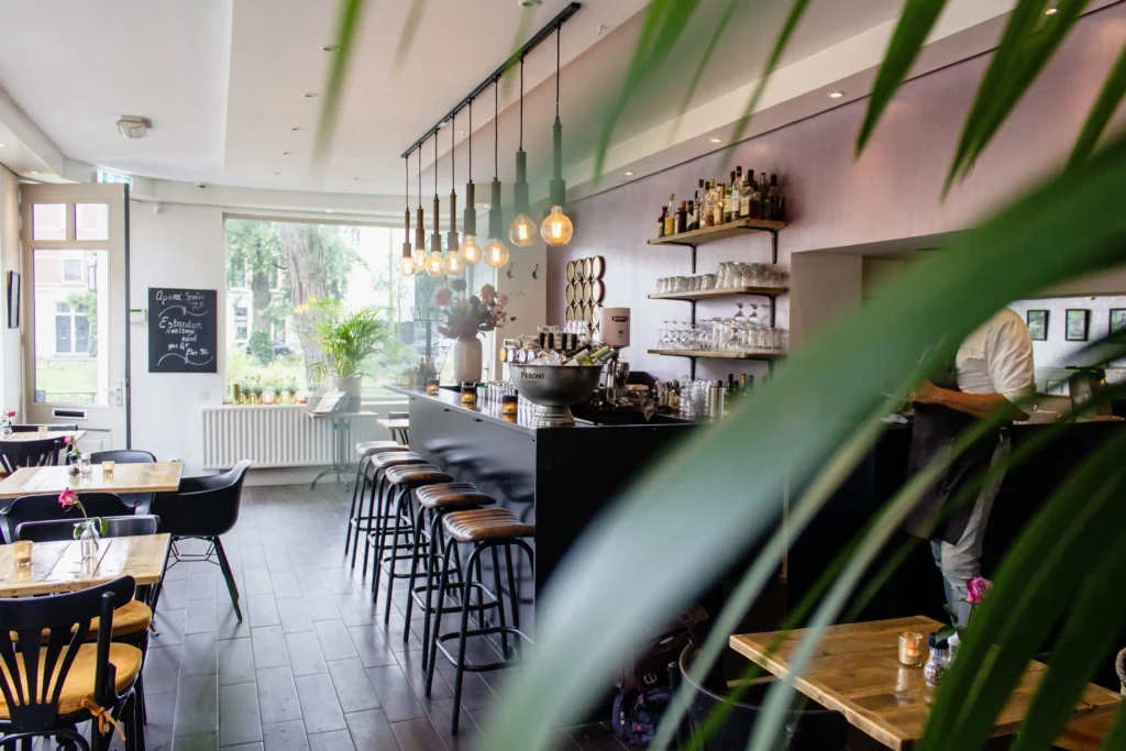 Bright modern café interior with a black bar counter, leather bar stools, Edison pendant lights, indoor plants and wooden tables in a relaxed bistro setting