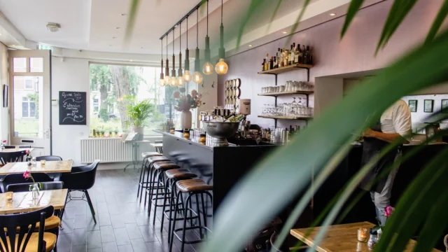Bright modern café interior with a black bar counter, leather bar stools, Edison pendant lights, indoor plants and wooden tables in a relaxed bistro setting
