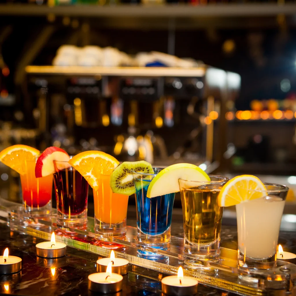 Row of colorful cocktail shots garnished with orange, kiwi and lemon slices on a bar counter with glowing tea lights in a warm night atmosphere