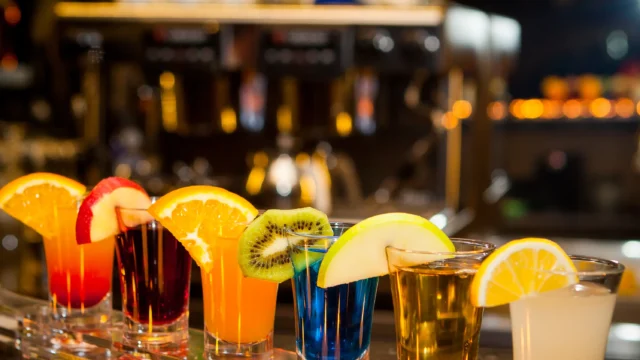 Row of colorful cocktail shots garnished with orange, kiwi and lemon slices on a bar counter with glowing tea lights in a warm night atmosphere