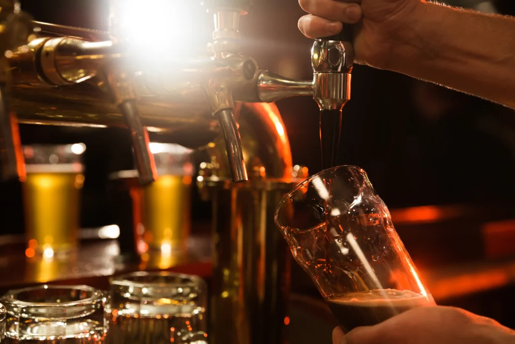 Bartender pouring draft beer from a chrome tap into a glass in a warmly lit bar with golden ambient lighting