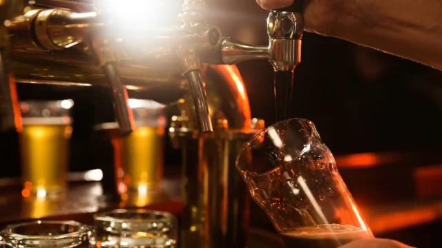 Bartender pouring draft beer from a chrome tap into a glass in a warmly lit bar with golden ambient lighting