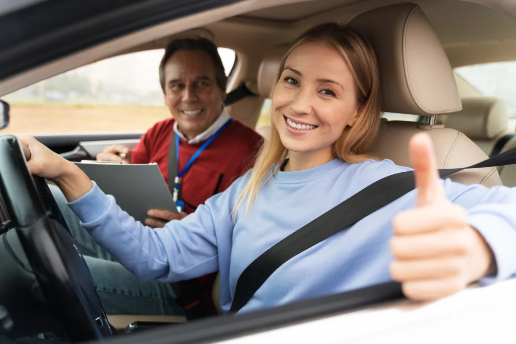 Smiling young woman giving thumbs up at the wheel during a driving lesson with an instructor holding a clipboard in the passenger seat