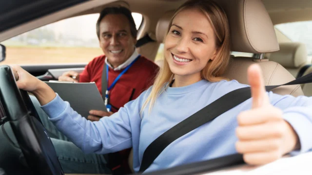 Smiling young woman giving thumbs up at the wheel during a driving lesson with an instructor holding a clipboard in the passenger seat