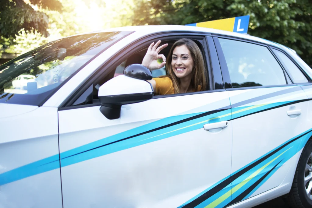 Smiling young woman giving an OK sign from the window of a learner driving school car with an L plate in sunny weather