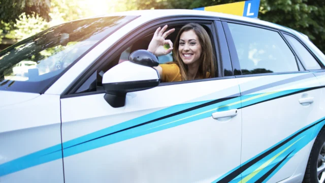 Smiling young woman giving an OK sign from the window of a learner driving school car with an L plate in sunny weather