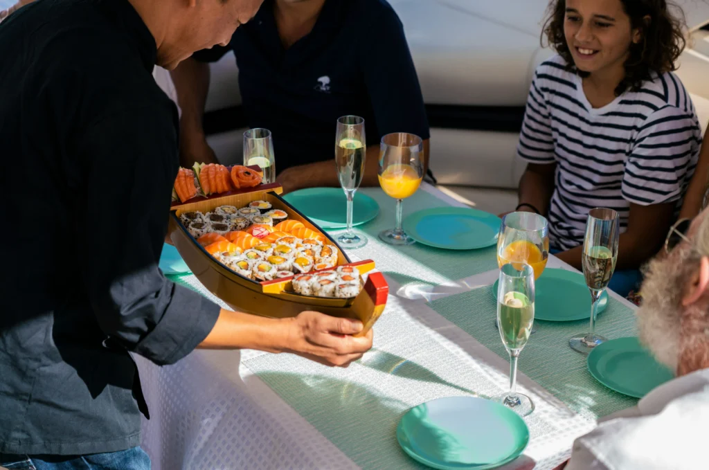Crew member serving a boat-shaped sushi platter to guests at a sunlit table on a luxury yacht with champagne glasses and turquoise plates