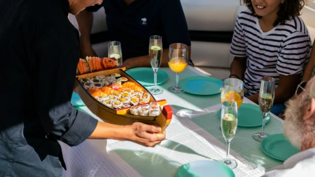 Crew member serving a boat-shaped sushi platter to guests at a sunlit table on a luxury yacht with champagne glasses and turquoise plates