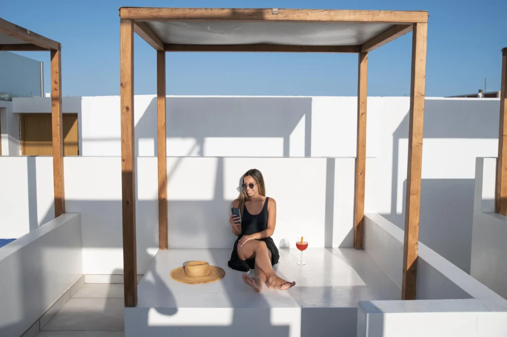 Woman in sunglasses sitting relaxed on a white rooftop terrace under a wooden pergola frame with a cocktail and straw hat on a sunny Greek island