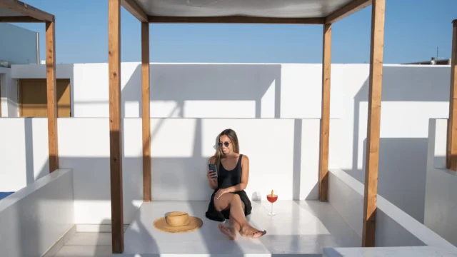 Woman in sunglasses sitting relaxed on a white rooftop terrace under a wooden pergola frame with a cocktail and straw hat on a sunny Greek island
