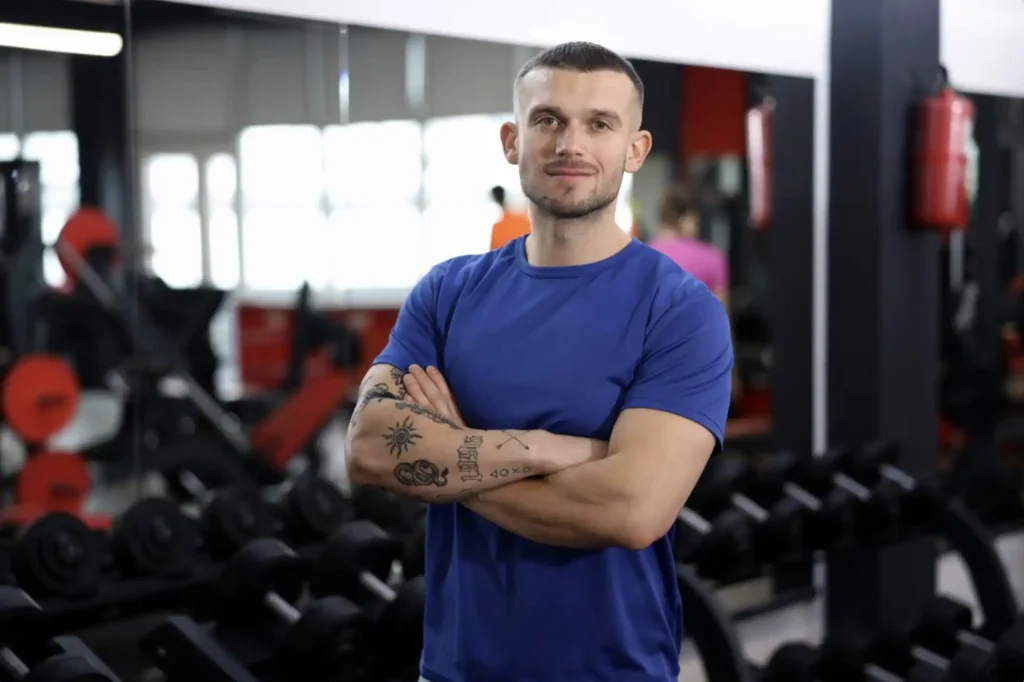 Confident personal trainer in a blue t-shirt with tattoos standing arms crossed in front of a dumbbell rack in a gym