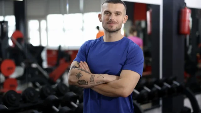 Confident personal trainer in a blue t-shirt with tattoos standing arms crossed in front of a dumbbell rack in a gym