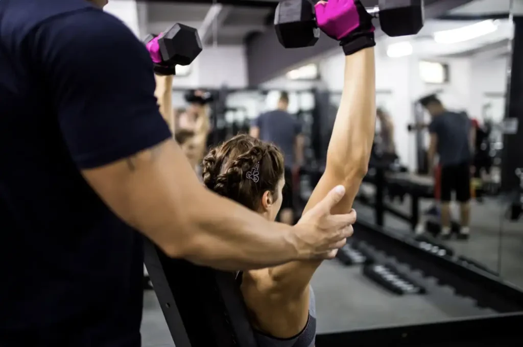 Personal trainer assisting a woman performing dumbbell shoulder presses on an incline bench in a gym with mirrored walls
