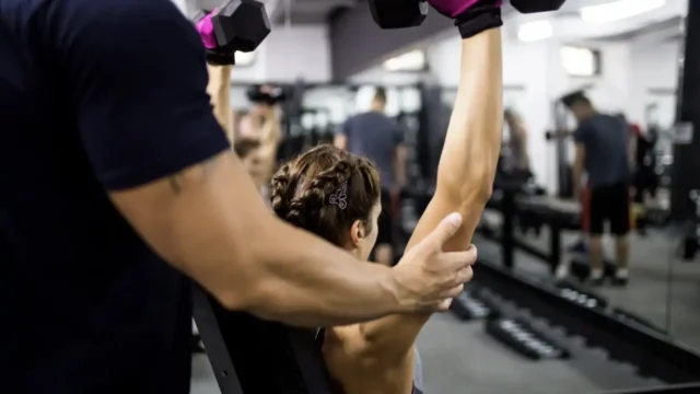 Personal trainer assisting a woman performing dumbbell shoulder presses on an incline bench in a gym with mirrored walls