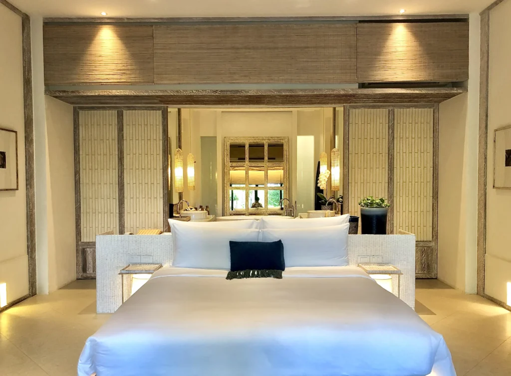 Elegant hotel bedroom with white king bed, bamboo wall panels and an open ensuite bathroom with a freestanding tub and warm ambient lighting