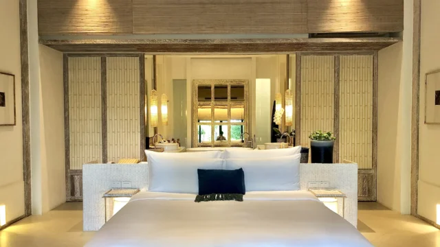 Elegant hotel bedroom with white king bed, bamboo wall panels and an open ensuite bathroom with a freestanding tub and warm ambient lighting