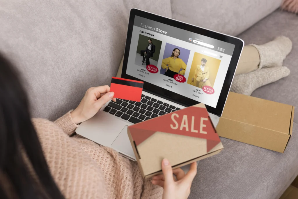 Woman browsing a fashion store website on a laptop while holding a credit card and a sale package box, sitting on a couch