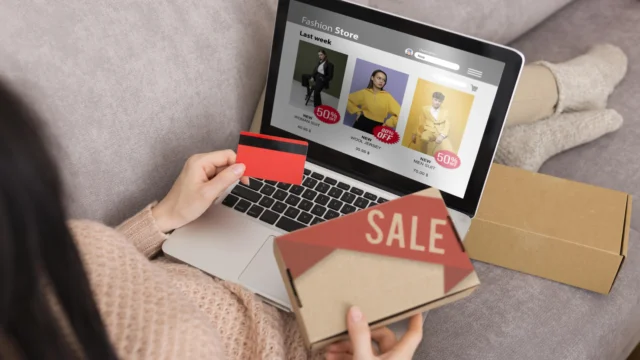 Woman browsing a fashion store website on a laptop while holding a credit card and a sale package box, sitting on a couch