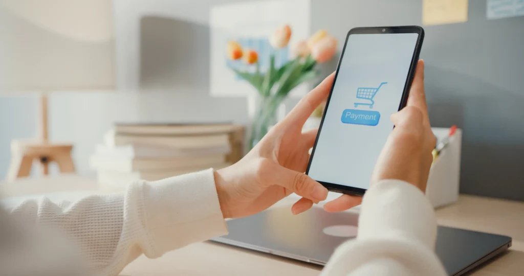 Woman tapping the payment button on a shopping app on her smartphone, sitting at a bright home desk with a laptop