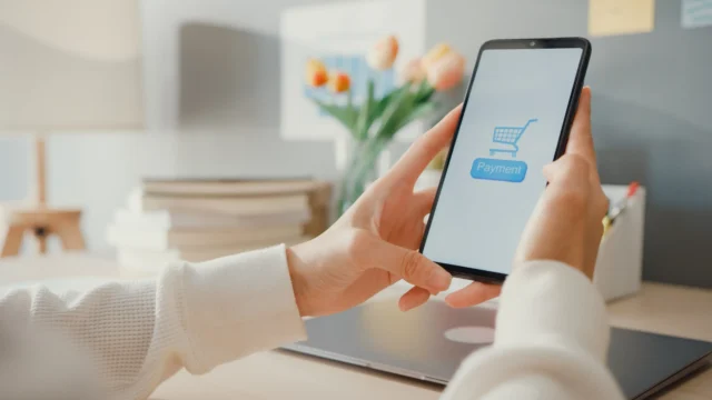 Woman tapping the payment button on a shopping app on her smartphone, sitting at a bright home desk with a laptop