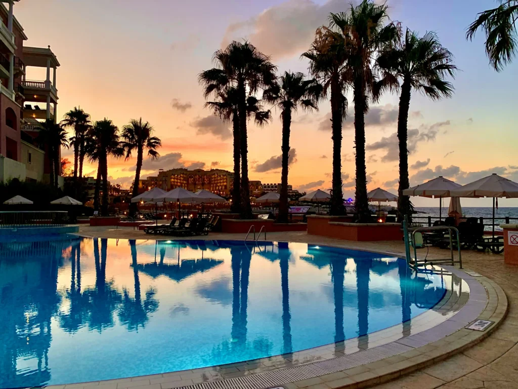 Illuminated hotel pool at sunset with palm tree silhouettes reflecting in the water and a warm orange sky over the sea