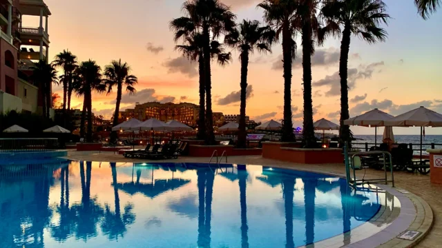 Illuminated hotel pool at sunset with palm tree silhouettes reflecting in the water and a warm orange sky over the sea