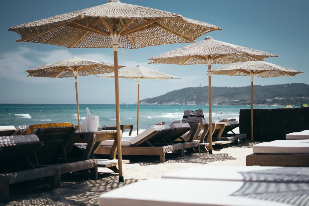 Exclusive beach club with woven rattan parasols and wooden sun loungers on white sand overlooking a turquoise sea with a green hillside in the background